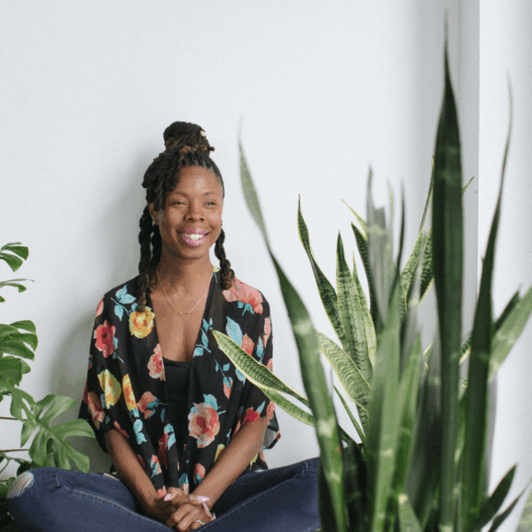 A serene portrait of BrownSkinned Plant Mama seated behind a lush snake plant, embodying plant-based healing and joy.